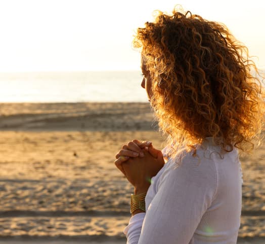 Anne-Marieke Visser aan zee bij zonsondergang, handen gevouwen, in gedachten.