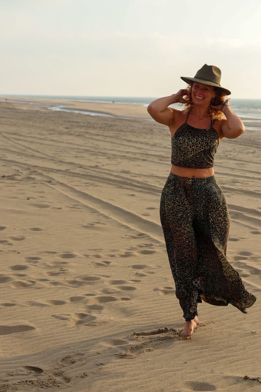 Anne-Marieke Visser loopt lachend over het strand bij zonsondergang, hoed op, armen omhoog.