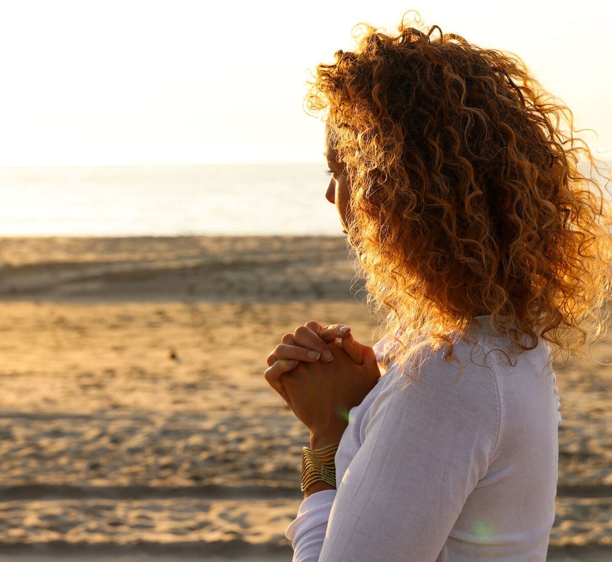 Anne-Marieke van opzij op het strand, handen gevouwen, omgeven door gouden avondlicht.
