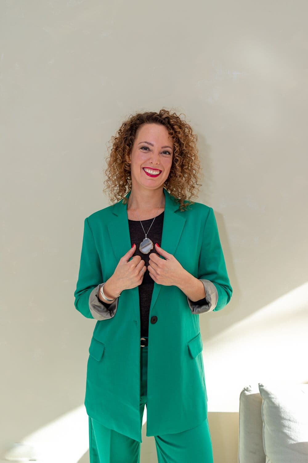 Anne-Marieke in groen pak, zelfverzekerd glimlachend tegen een lichte muur.