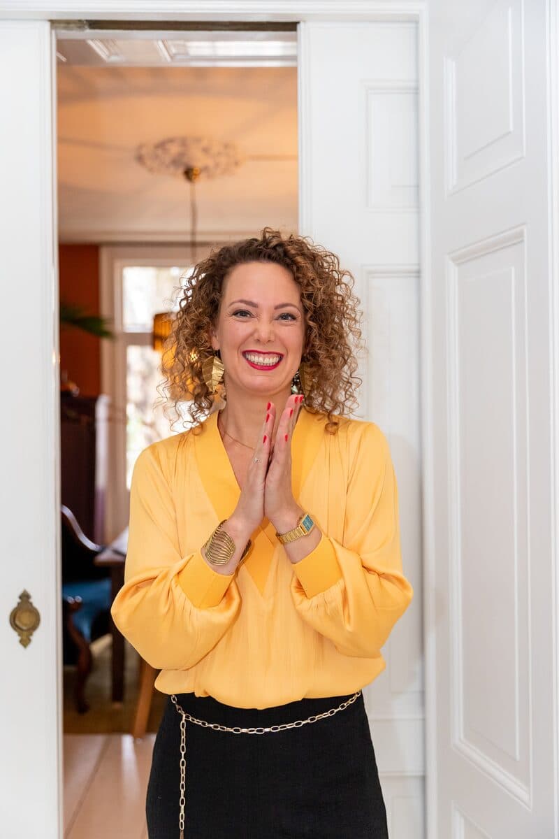 Anne-Marieke Visser in gele blouse, breed lachend vanuit een deuropening.